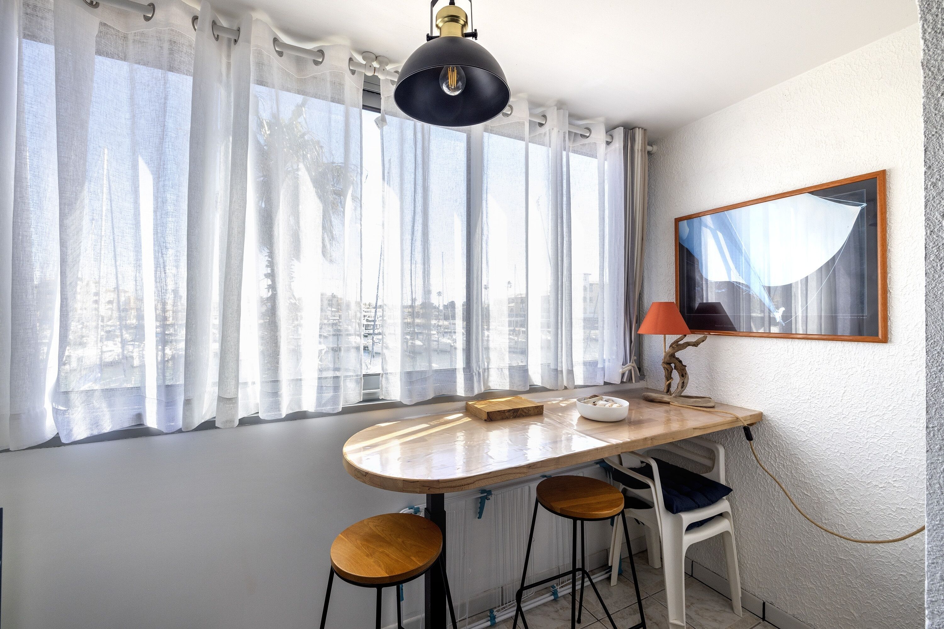 A dining area with a wooden table, two stools, a lamp, and a cityscape view through sheer curtains.