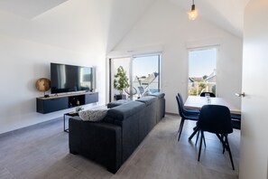 Ein modernes Wohnzimmer mit Fernseher, einer Couch, einem Esstisch mit Stühlen und großen Fenstern.