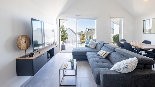 A modern living room with a large sectional sofa, a flat-screen TV, and a dining area with a wooden table and chairs. - L'Escale de Césarine - 1 km de la plage (Carnac)