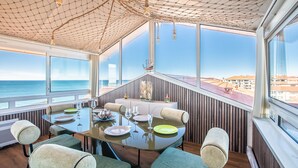 Uma sala de jantar com mesa posta para quatro pessoas, janelas amplas e vista para o mar.
