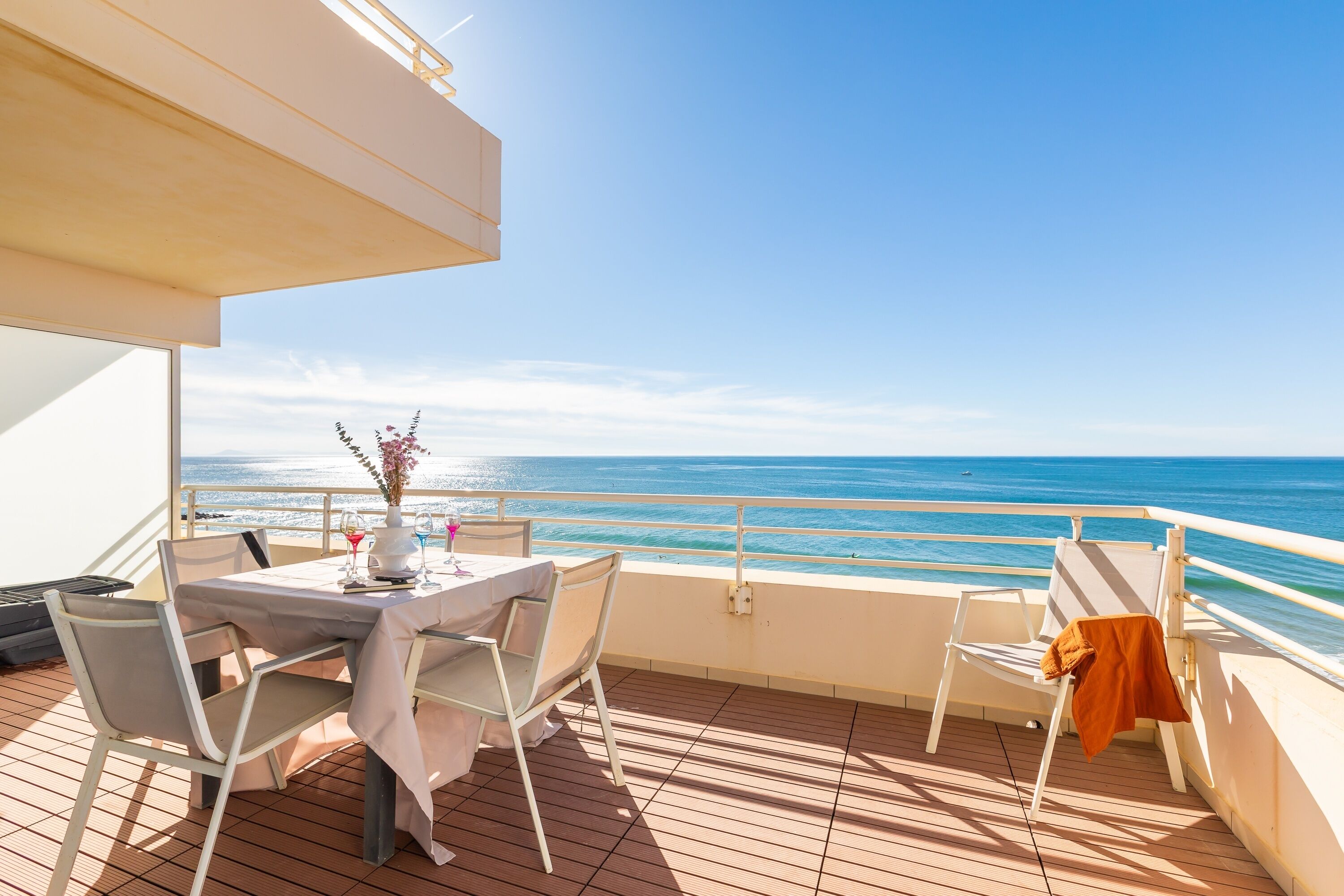 Un balcon avec une table et des chaises, un vase rempli de fleurs et une vue sur la mer.