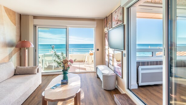 Uma sala de estar moderna com sofá, mesa de centro e obras de arte, que dá para uma varanda com vista para o mar.