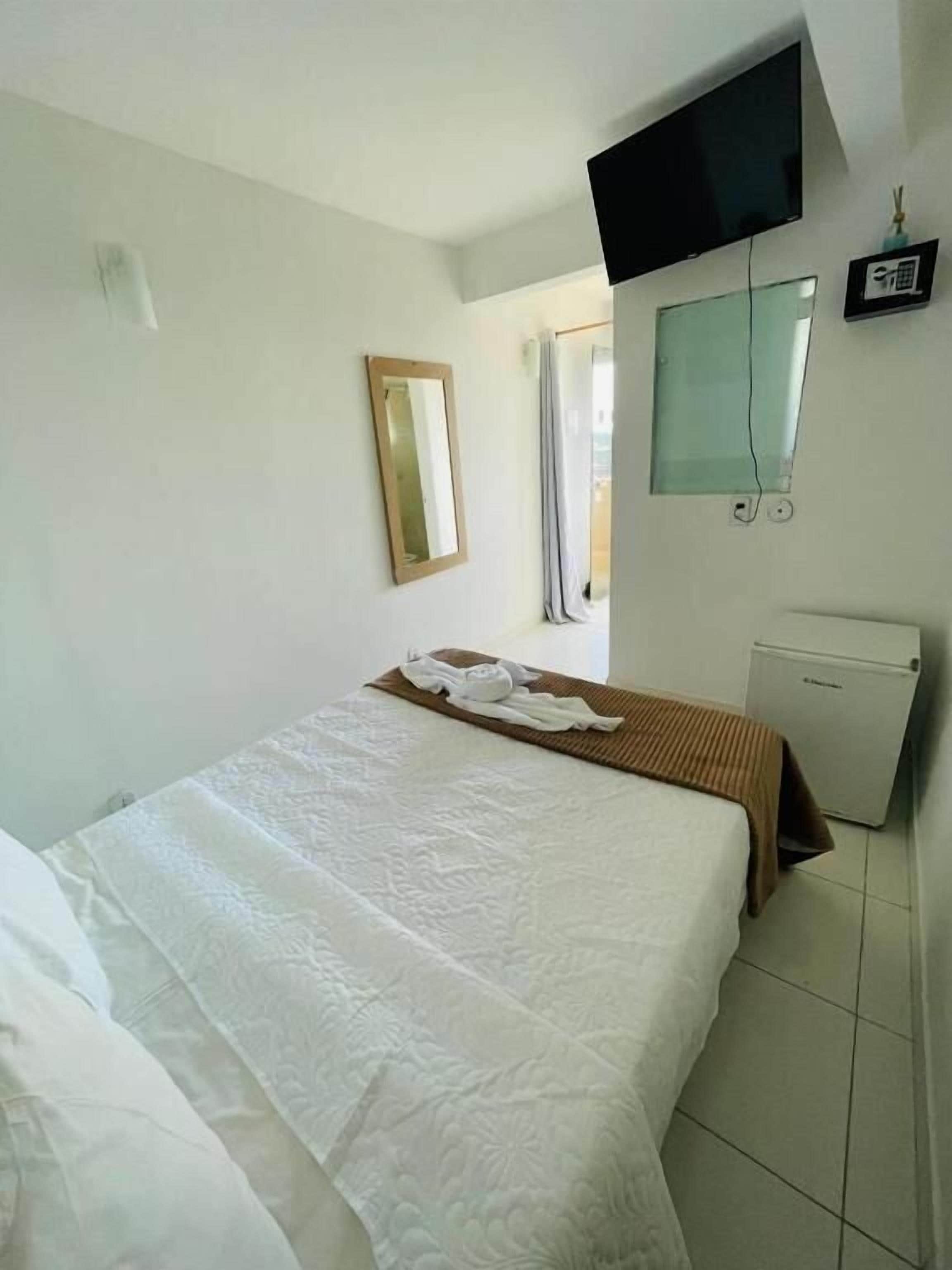 A hotel room with a bed, a flat-screen TV, a small refrigerator, and a window with curtains.