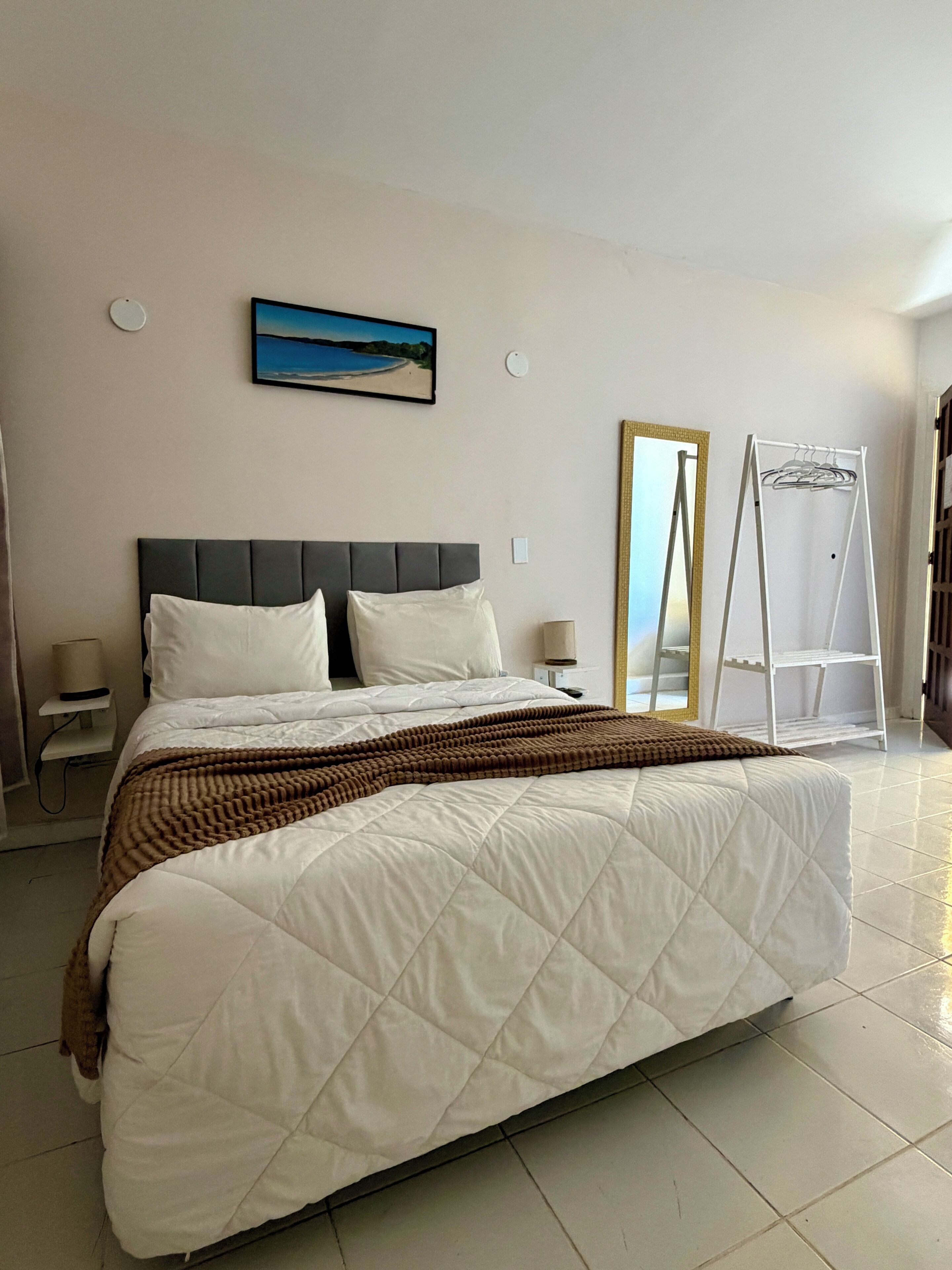 A hotel room with a large bed, a bedside table with a lamp, a painting on the wall, and a white wardrobe with a mirror.