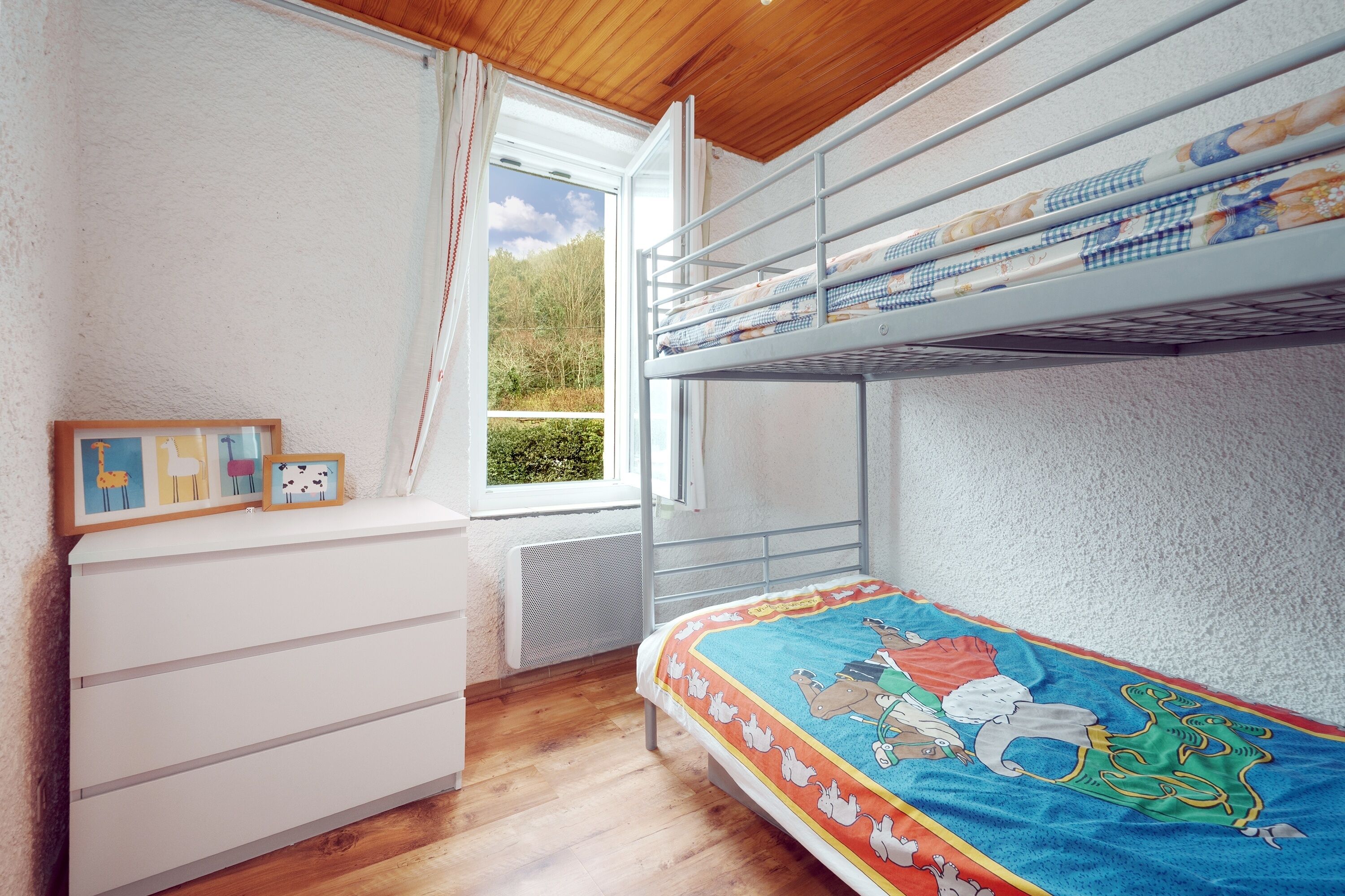 A bunk bed with a colorful bedspread, a white dresser, and a window with curtains.
