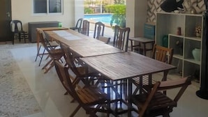 A dining area with a long wooden table and chairs, a TV mounted on the wall, and a view of a swimming pool outside. - Elicon Suites  (Baclayon)