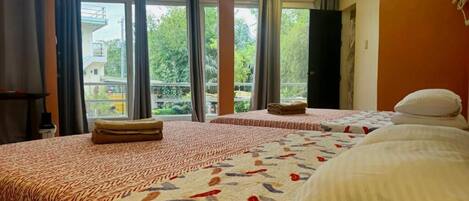 A hotel room with two beds, a window with curtains, a view of greenery outside, and a door to the outside.