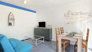 Uma sala de estar moderna com um sofá azul, uma mesa de jantar com tampo de vidro e uma televisão sobre um móvel.