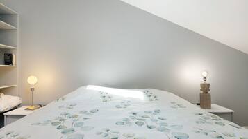Een slaapkamer met een schuine plafond, een bed met een deken met bladmotief, een bedlampje en een wandlamp.