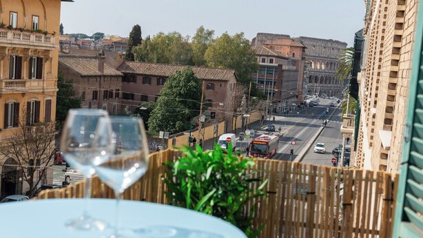 A table with a glass of wine, a view of a city street with buildings and a bus, and a wooden fence.