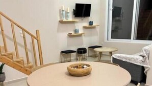 Spiseplads med et rundt bord, fire træstole, en skål med snacks, et fjernsyn på væggen og en trappe.
