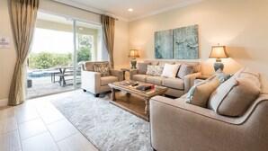A living room with a sofa, armchair, coffee table, and two lamps. There is a painting on the wall and a window with curtains. - 6BR Home w/ Pool, Theater & Game Room (Kissimmee)
