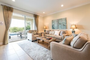 Ein Wohnzimmer mit einer Couch, einem Sessel, einem Couchtisch und zwei Lampen. An der Wand hängt ein Gemälde, und ein Fenster ist mit Vorhängen drapiert.