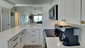 A modern kitchen with white cabinetry, a built-in microwave, a coffee machine, and a dining area with a view. - Pointe South 302 | Gulfside Escape + Resort Pool (Fort Myers Beach)