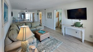 A living room with a sofa, a coffee table, a TV, and a dining area in the background. - Pointe South 302 | Gulfside Escape + Resort Pool (Fort Myers Beach)