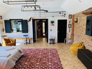Um quarto espaçoso com uma cama grande, um tapete vermelho estampado, um pufe amarelo, uma parede de tijolos e uma luminária pendente.