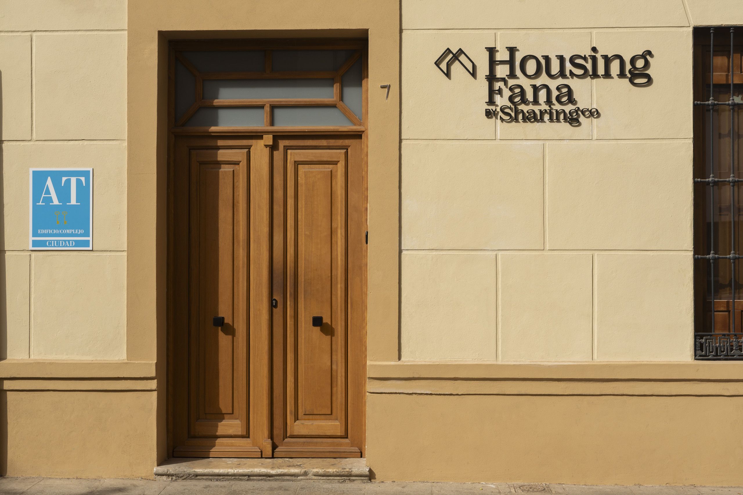 A building entrance with a wooden double door, a sign for 'Housing Fana Sharingco,' and a blue sign with 'AT CIUDAD' on the left.
