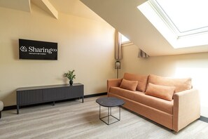 Ein modernes Wohnzimmer mit einer Couch, einem kleinen runden Tisch, einer Topfpflanze, einer schwarzen Konsole und einem Schild an der Wand.