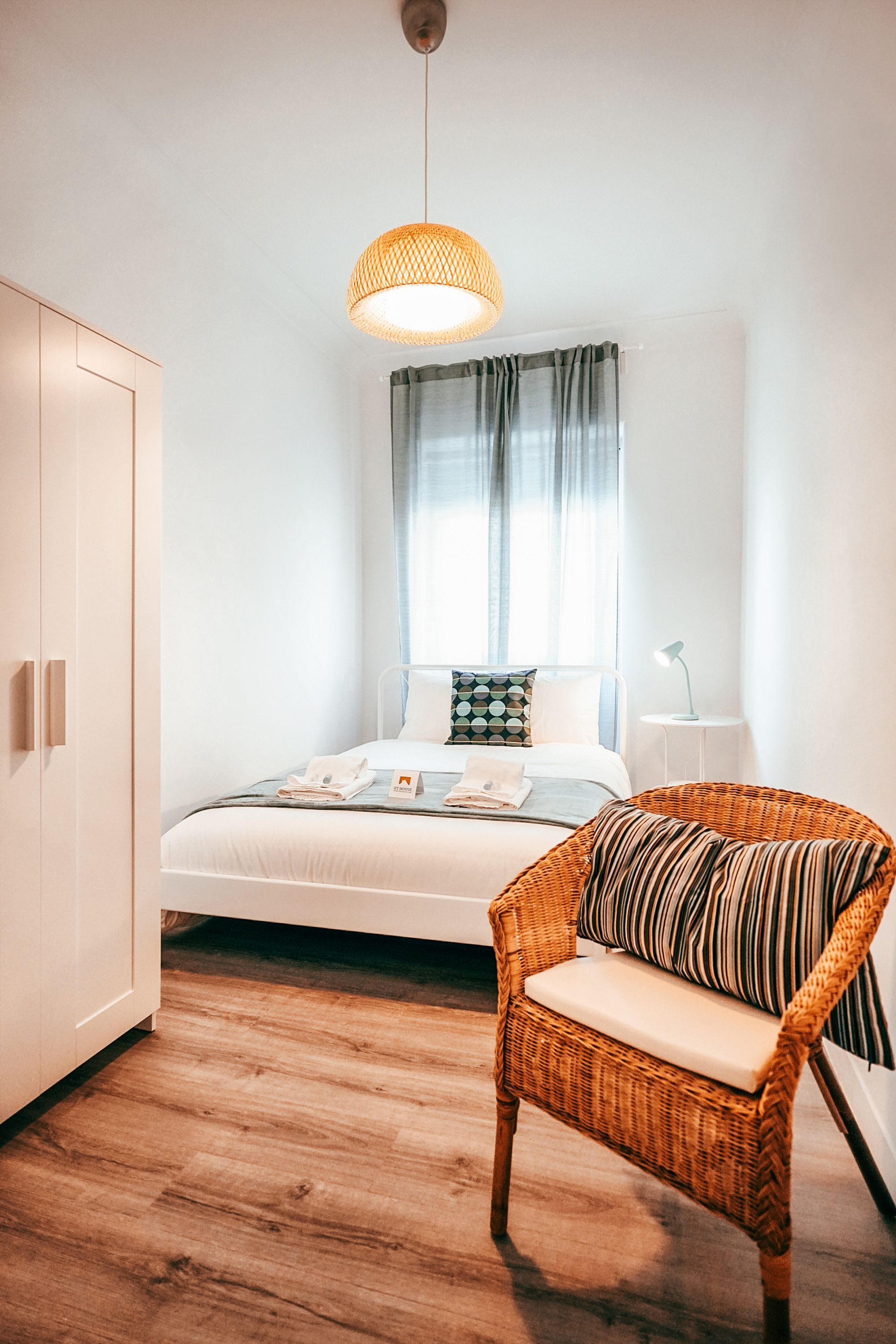 Une chambre à coucher comprenant un lit, une chaise en osier, une table de chevet et un luminaire suspendu.