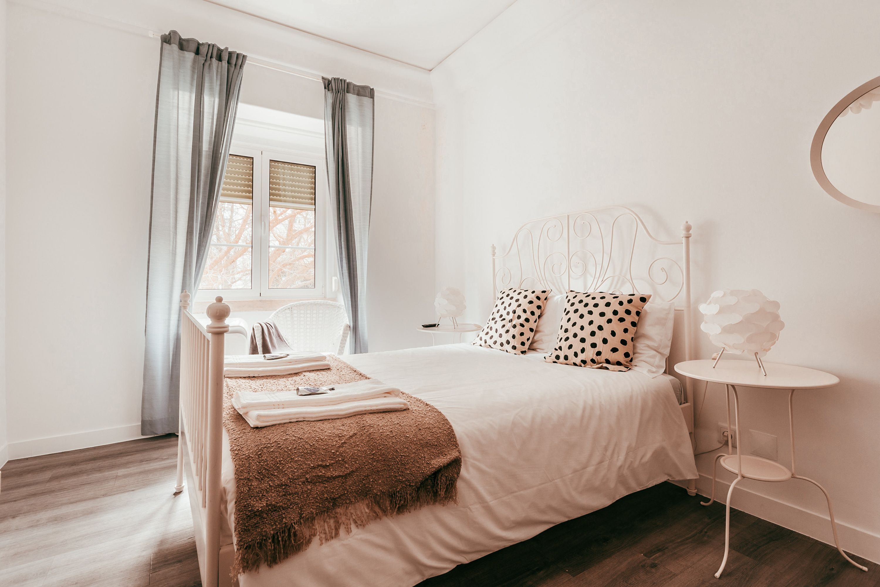 A bedroom with a bed, a bedside table, a round mirror, and a window with curtains.
