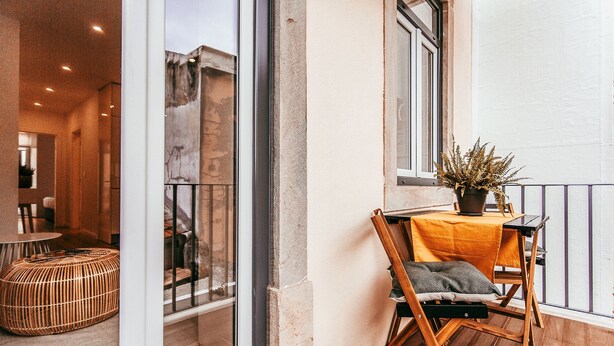 A balcony with a wooden chair, a small table with a plant, and a wicker ottoman. - Prestige Apartment By Gt House (Lisbon)