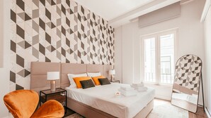 A modern hotel room with a geometric-patterned wall, a large bed with white linens, a side table with a lamp, a chair, and a mirror. - Elegant Sophisticated Campo Ourique by GT House (Lisbon)