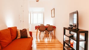Ein Wohnzimmer mit einer Couch, einem Esstisch mit Stühlen, einem Fernseher auf einem Schrank und einer Pendelleuchte.