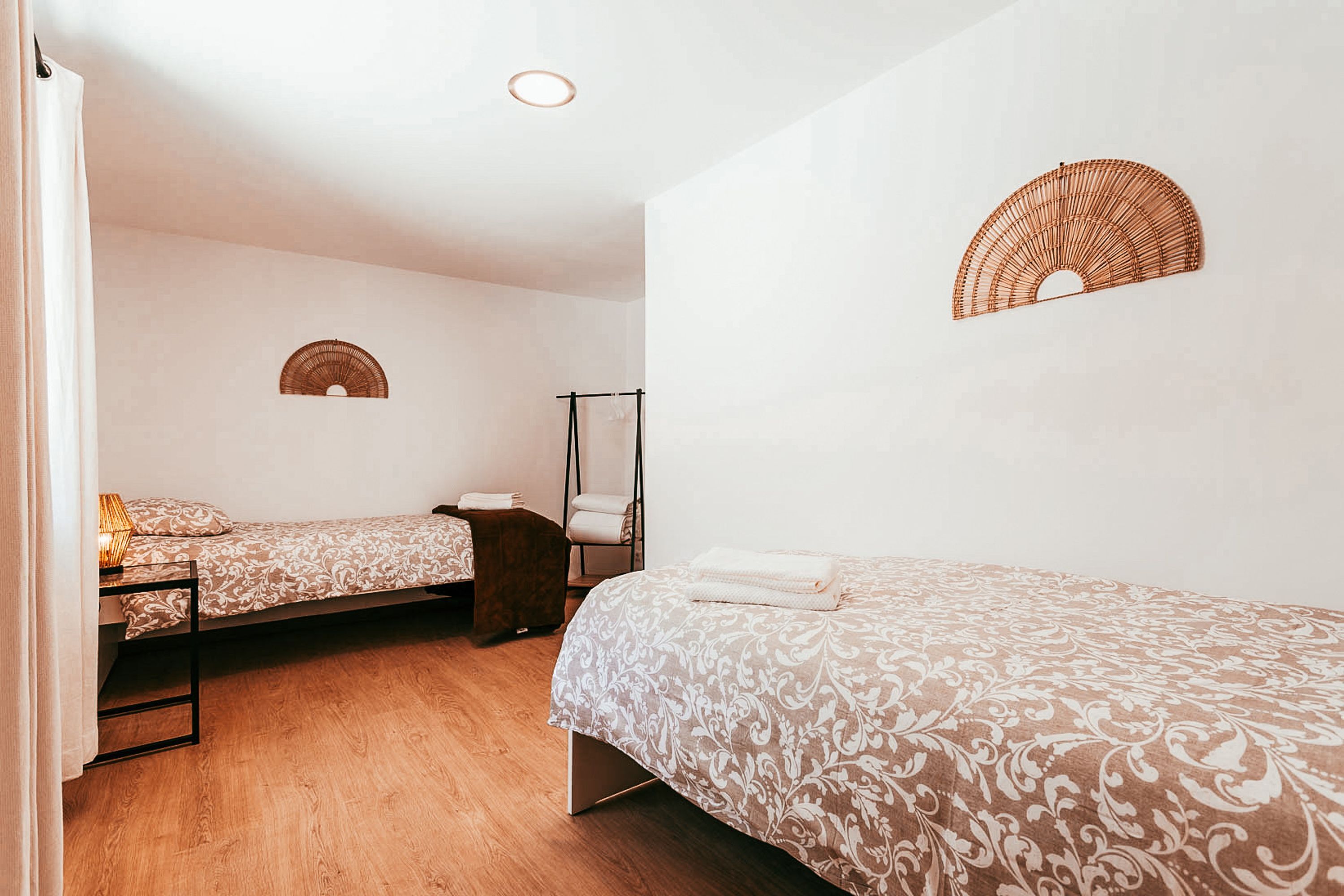 Een slaapkamer met twee bedden, een houten vloer, een staande ventilator en een ventilator aan de muur.