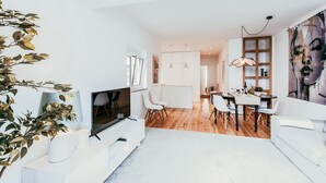 Una sala de estar moderna con un televisor de pantalla plana, muebles blancos, un comedor con mesa de madera y un cuadro abstracto de gran tamaño.