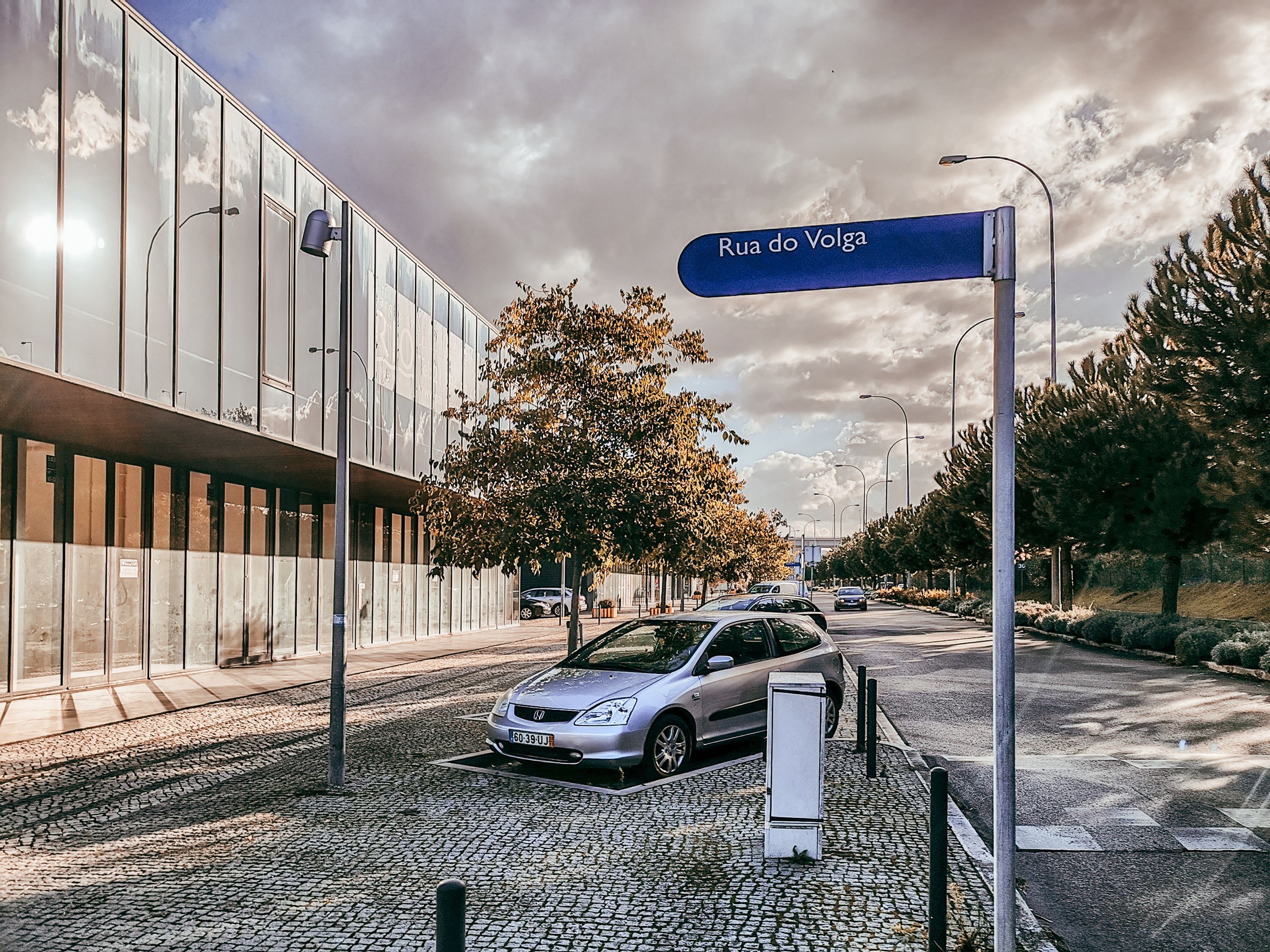 Straßenansicht mit einem Schild, das 'Rua do Volga' zeigt, ein modernes Gebäude mit Glasfassade, ein am Straßenrand geparktes Auto und eine gepflasterte Straße.
