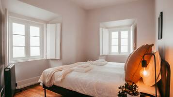 Un dormitorio con una cama, dos ventanas, un radiador, una planta en maceta y un aplique de pared.