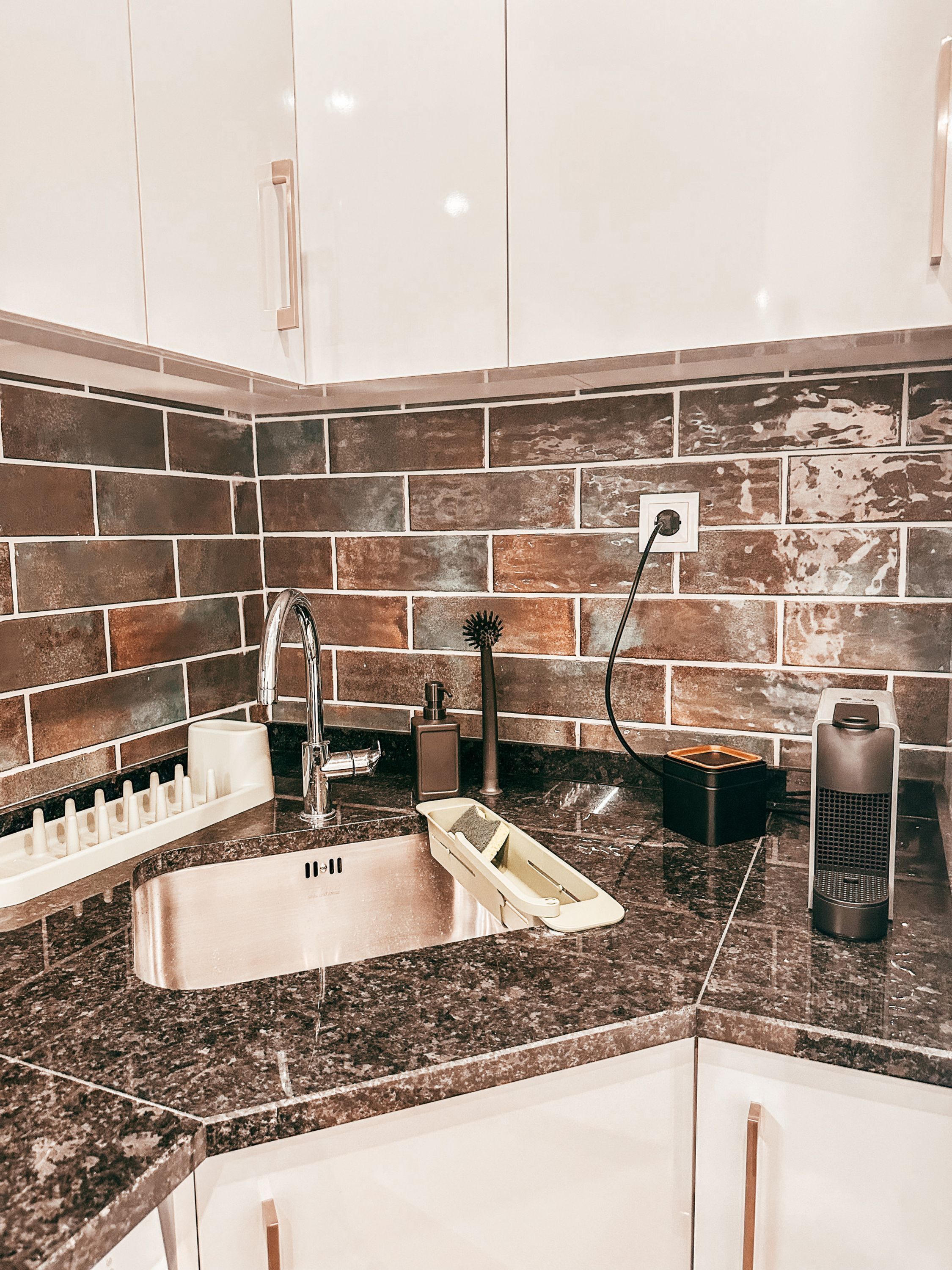 A kitchen with a stainless steel sink, a built-in soap dispenser, a coffee maker, and a brick-patterned backsplash.