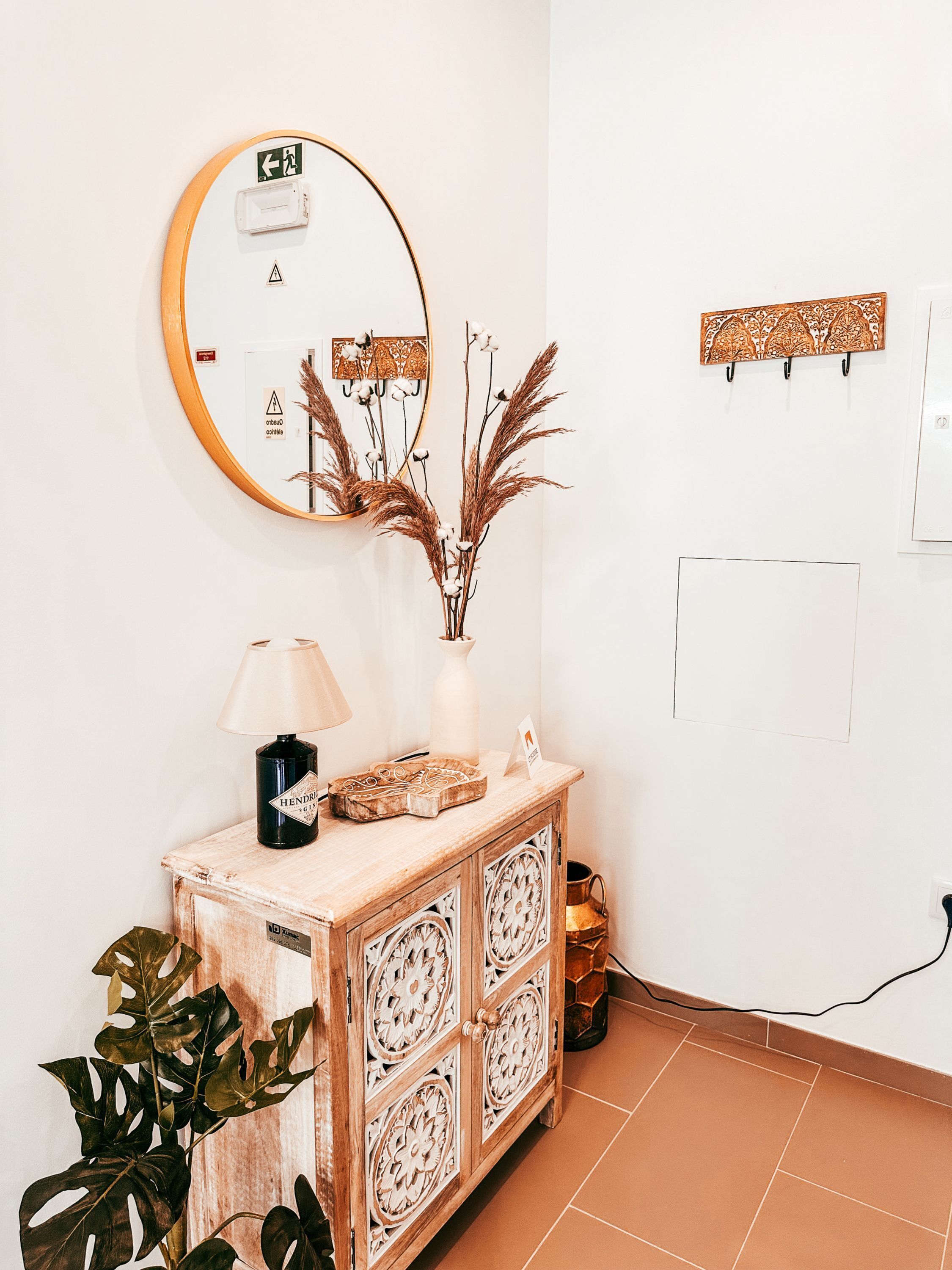 A room with a round mirror, a wooden cabinet with decorative panels, a vase with dried plants, a lamp, and a potted plant.