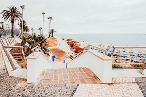 Un hotel frente a la playa con una escalera que da a la arena, mobiliario exterior y sombrillas.