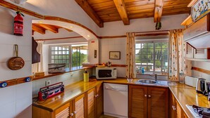 A compact kitchen with wooden cabinets, a microwave, a sink, and a window with curtains. - Jacarand Apartment in Quinta Velha by GT House (Portimão)