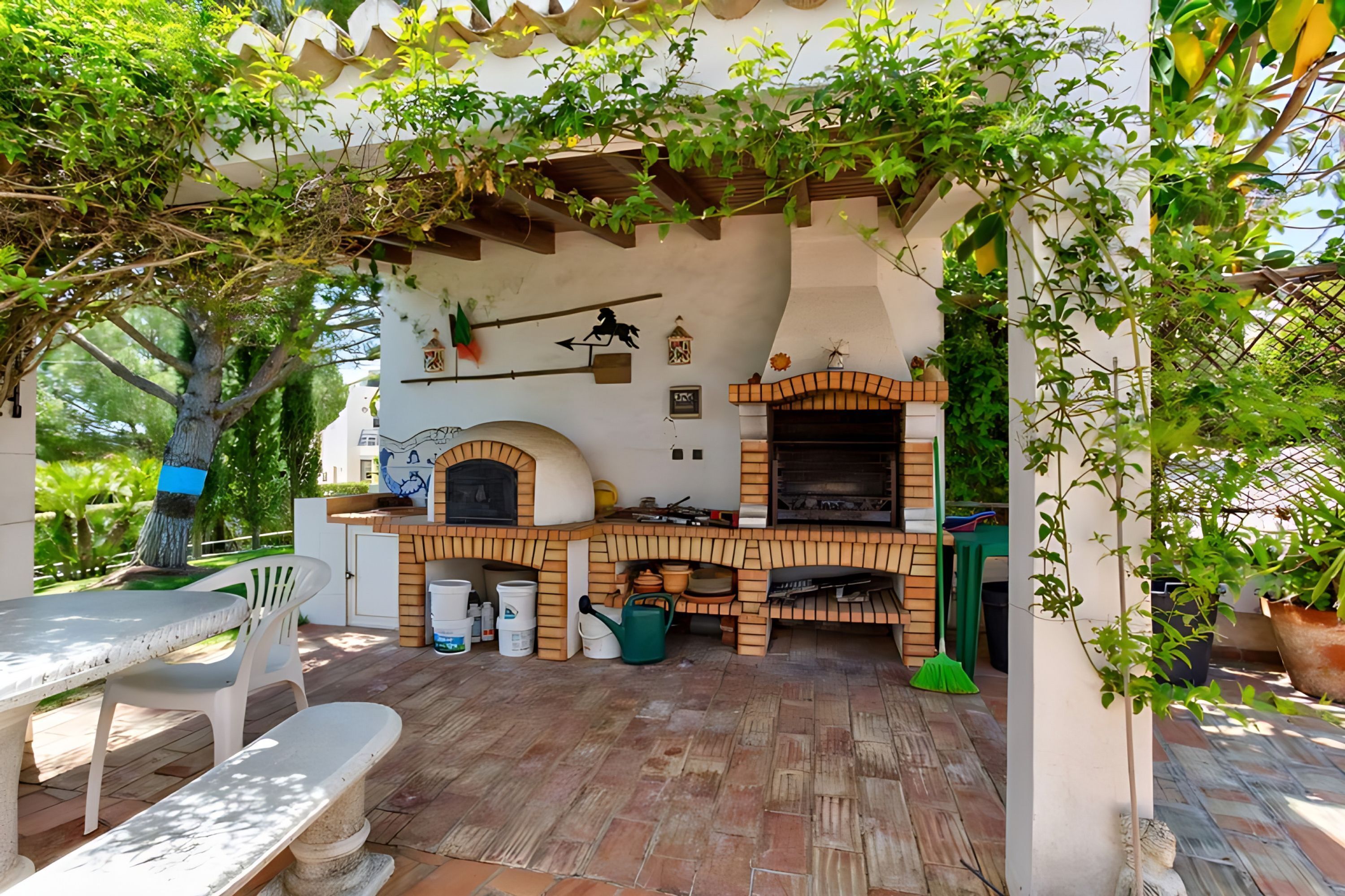 Een buitenkeuken met een bakoven, een tafel en stoelen, omgeven door potplanten en een pergola.