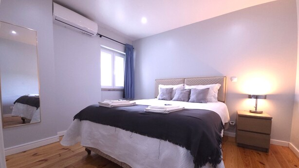 A bedroom with a bed, a bedside table with a lamp, a window with curtains, and an air conditioning unit. - Sesimbra Terrace Escape By Gt House (Sesimbra)