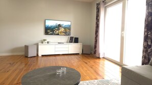 A living room with a flat-screen TV mounted on the wall, a white entertainment unit, a round coffee table, and a sliding glass door leading to a balcony. - Sesimbra Terrace Escape By Gt House (Sesimbra)