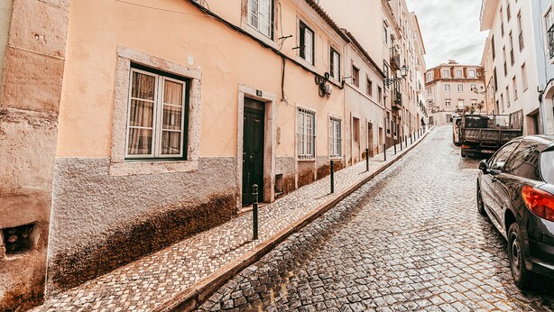 Uma rua de paralelepípedos flanqueada por edifícios, um carro estacionado e um caminhão ao longe.