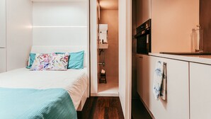 Quarto de hotel pequeno, com uma cama, um banquinho, uma pia e um espelho em espaço separado.