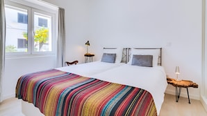 Ein Schlafzimmer mit einem großen Bett, einem Fenster mit Vorhängen, einem Nachttisch mit Lampe und Blick auf Gebäude.