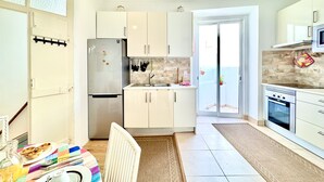 Eine moderne Küche mit Kühlschrank, Herd und Spüle. Ein Esstisch mit bunten Tischdecken und ein Stuhl sind ebenfalls vorhanden.