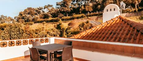 A terrace with a table and chairs, a tiled roof, and a view of trees and hills.