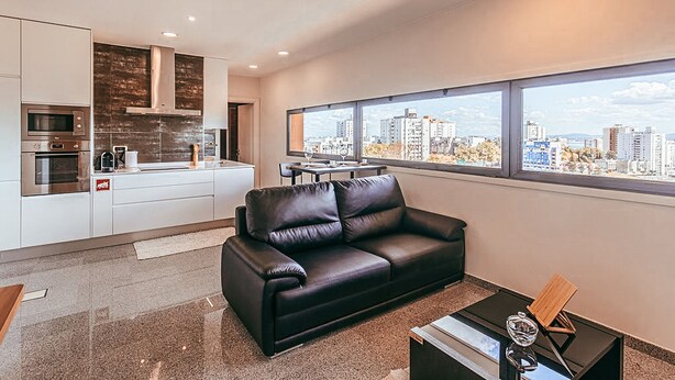 A modern kitchen with built-in appliances and a dining area. A black leather sofa is positioned in front of large windows offering a city view. - Trendy Apartment River View By Gthouse (Lisbon)