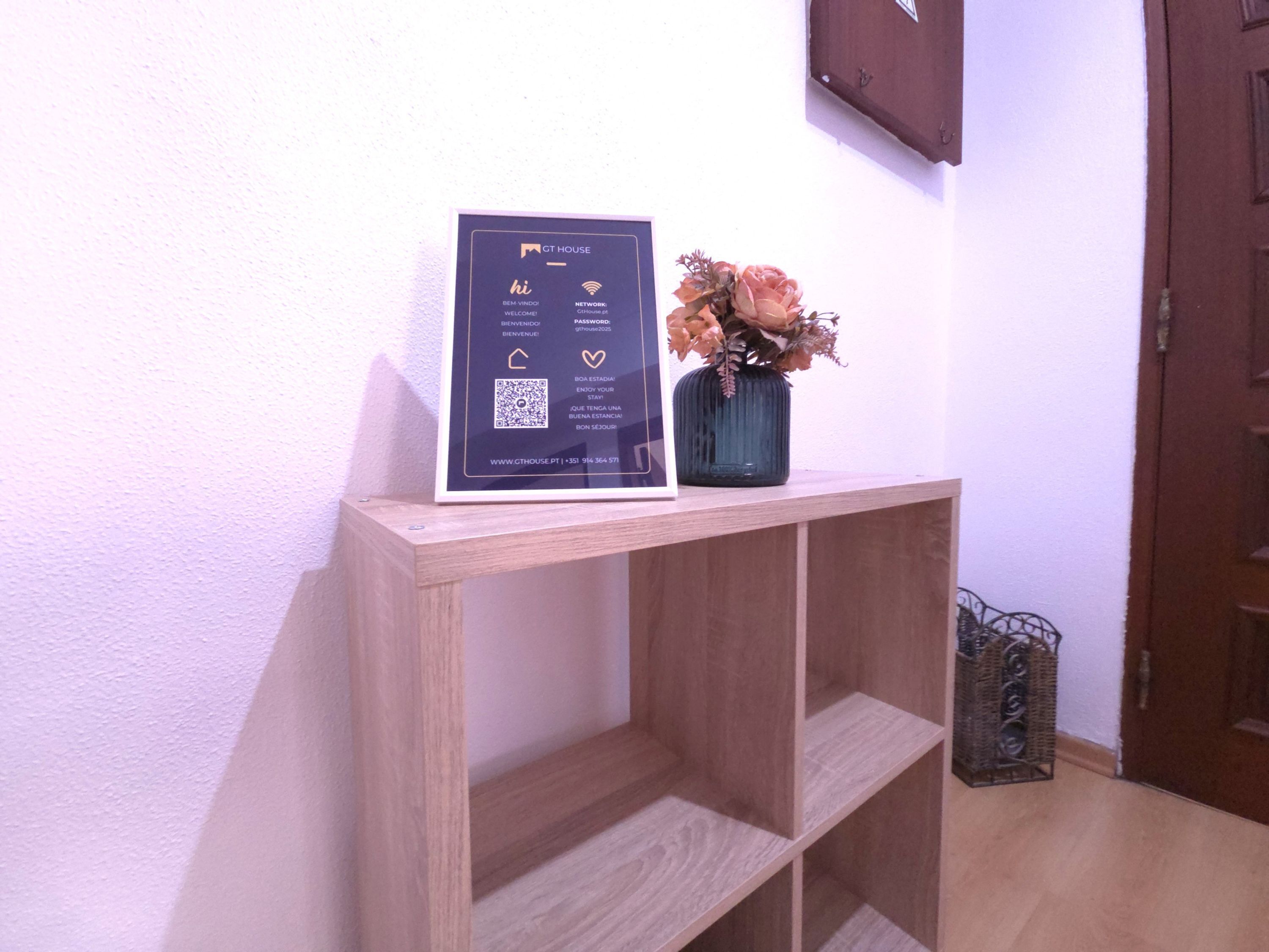 A wooden console table with a framed welcome sign, a QR code, and a vase with dried flowers.