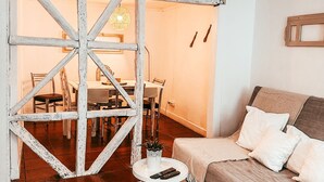 Una habitación con un separador de madera rústico, un sofá con almohadones, una mesita redonda pequeña con un control remoto y una lámpara colgante.