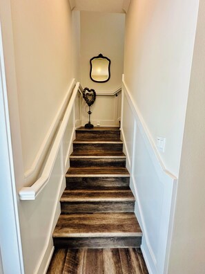 Un escalier avec des marches en bois et des murs blancs, menant à un palier agrémenté d’un miroir et d’un objet décoratif en forme de cœur.