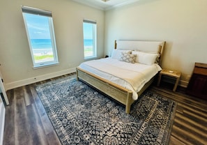 Une chambre avec un grand lit, deux fenêtres, un tapis à motifs et une vue sur l’océan.