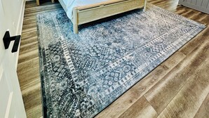 Un dormitorio con cama, ventilador de techo, mesita de noche y suelo de madera con alfombra estampada.