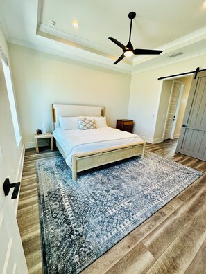 A bedroom with a bed, a ceiling fan, a nightstand, a bedside table, a patterned rug, and a wooden floor. - Beautiful Oceanfront House with Pool in Flagler Beach (Flagler Beach)